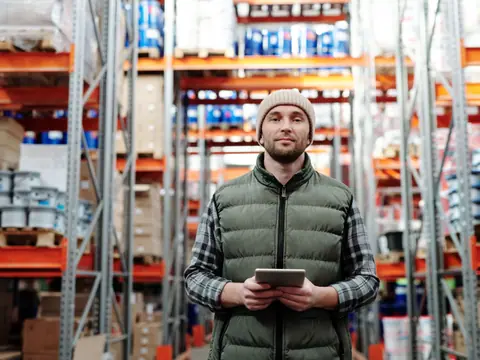 Warehouse worker using a tablet beside shelves of stored goods.